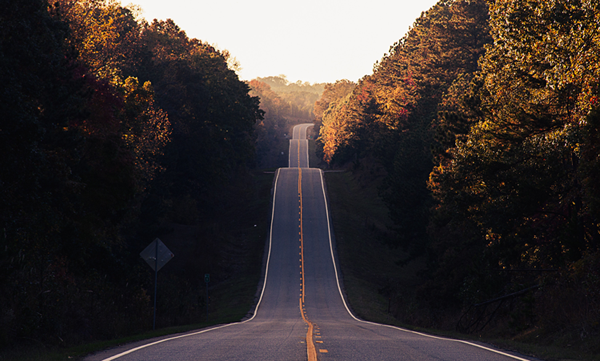 A long road stretches off into the distance, climbing over hills and cutting through a forest.